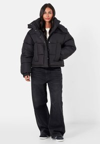 Kvinna med långt mörkt hår som bär en svart pufferjacka, svarta vida jeans och svarta sneakers, stående mot en enkel vit bakgrund.