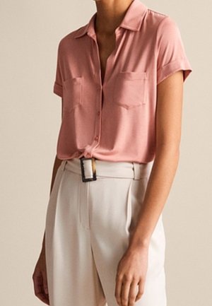 Kobieta w różowej koszuli na krótki rękaw z guzikami, włożonej do białych spodni z wysokim stanem, z czarną klamrą paska.