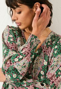 Blumenmuster Bluse mit langen Ärmeln in Grün, Kastanienbraun und Creme. Merkmale sind ein gesmoktes Handgelenkdetail und ein V-Ausschnitt, ergänzt durch kunstvolle botanische Muster.