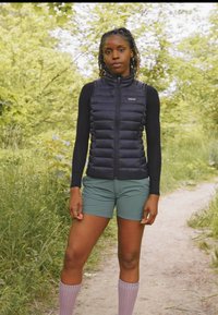 Svart pufferväst med vertikal sömnad, tillsammans med gröna shorts. Modellen bär knähöga pastell-lila strumpor och står på en stig bland grönska.