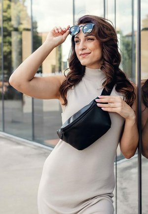 Schwarze Leder-Gürteltasche über ein hellbeiges Kleid getragen; Modell passt Sonnenbrille an, zeigt welliges Haar und ein elegantes, modernes Design.