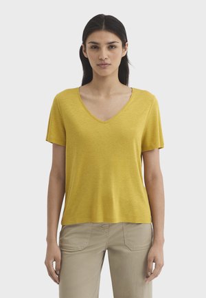 Donna con capelli lisci e scuri che indossa una maglietta gialla a scollo a V e maniche corte e pantaloni beige, in piedi contro uno sfondo chiaro e uniforme.