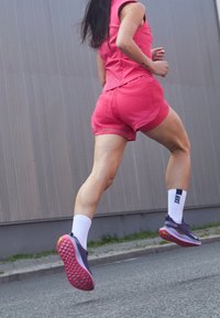 Top atletico rosa e pantaloncini, calze bianche e scarpe viola con suole texture, catturati mentre corrono su una superficie urbana.