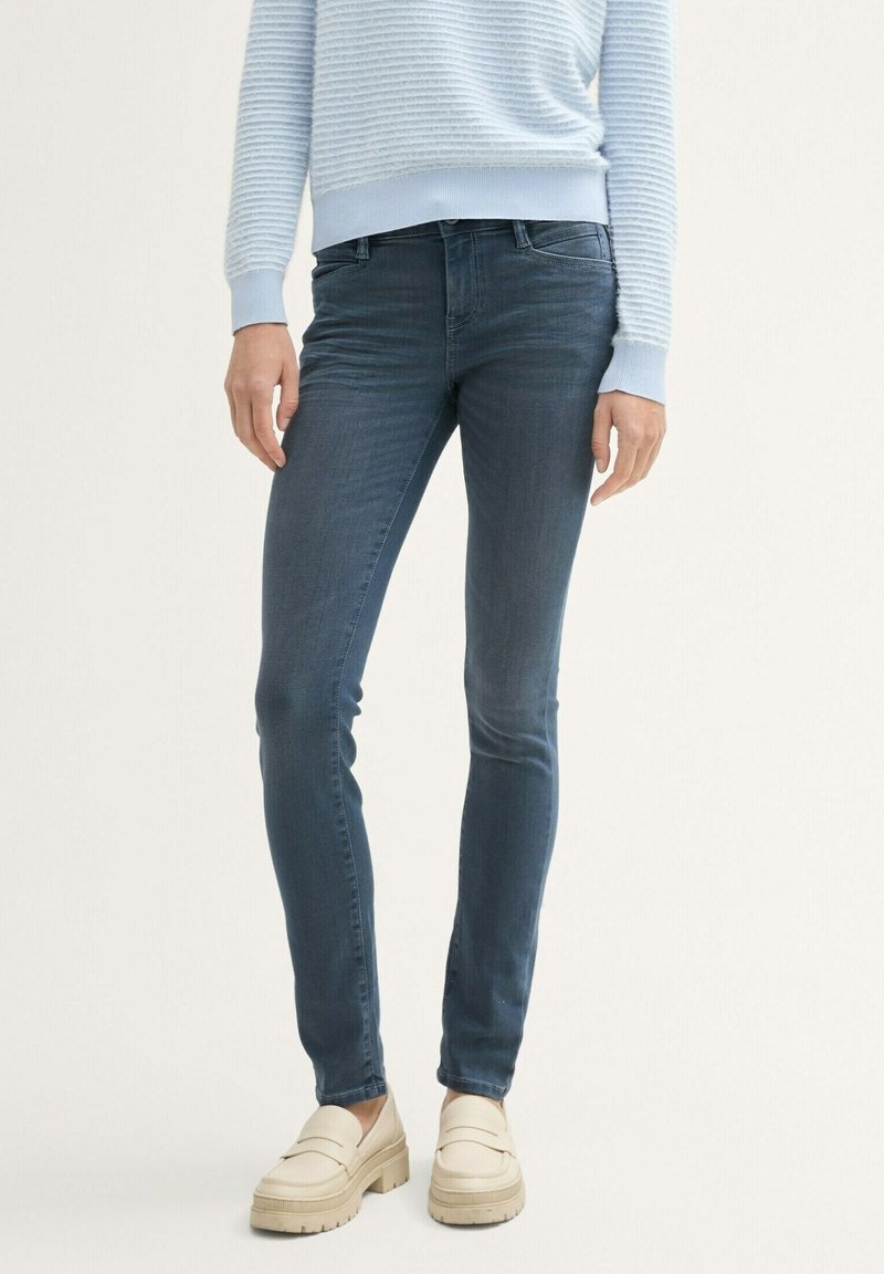 Personne portant un jean slim bleu foncé, un pull côtelé bleu clair et des mocassins beiges épais, debout devant un fond clair uni.