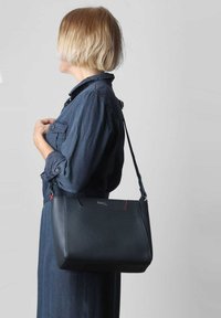 Zwarte leren handtas met een gestructureerde vorm, rode accentstiksels en een lange verstelbare schouderband, vastgehouden door een persoon in een blauw shirt.