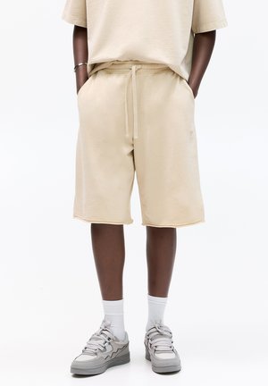 Shorts en coton beige avec une taille élastique et un cordon de serrage, dotés de poches latérales et de bords brut, associés à des baskets grises et des chaussettes blanches.