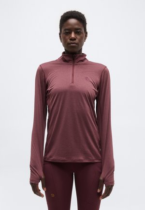 Γυναίκα που φοράει μακρυμάνικο μπλουζάκι με φερμουάρ σε μπορντό χρώμα και αντίστοιχο κολάν, στέκεται μπροστά σε απλό ανοιχτό φόντο.