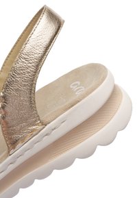 Detail zlatého koženého pásku na sandálu s polstrovanou béžovou semišovou stélkou a bílou texturovanou klínovou podrážkou.