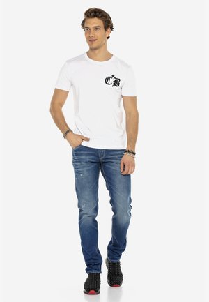 Weißes T-Shirt mit kurzen Ärmeln und schwarzem Logo auf der Brusttasche, kombiniert mit blauen, verwaschenen Jeans und schwarzen, mit Nieten versehenen Sneakers.
