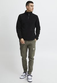 Sort fleece pullover med kvart lynlås, olivenfarvede cargo-bukser med sidelommer og hvide sneakers med marineblå detaljer, sat mod en ensfarvet baggrund.