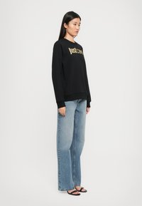 Donna in felpa nera "Justcavelli", jeans a gamba larga azzurri e sandali neri open-toe su sfondo bianco.