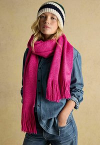 Jasně růžová pletená šála s třásněmi na koncích, nošená přes džínovou košili. Šála má texturované tkaní, které dodává hloubku jejímu designu.