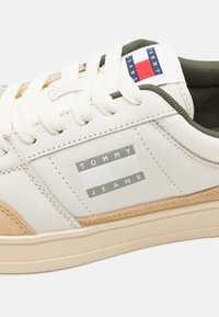 Sneaker bianca in pelle con accenti in suede color tan, suola in gomma beige e logo "TOMMY JEANS" grigio ben visibile sul lato. Lacci in cotone bianchi.