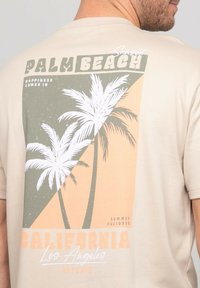 T-shirt beige avec un design graphique comprenant des palmiers, des formes géométriques orange et vertes, ainsi que le texte "Palm Beach California."