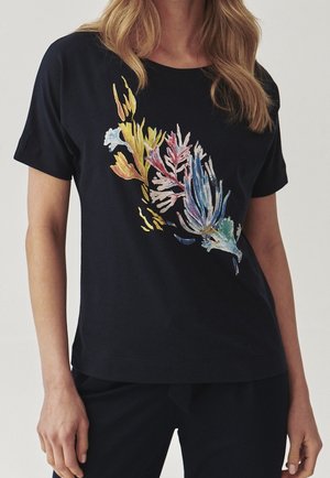 Kobieta ubraną w czarną koszulkę z krótkim rękawem z kolorowym abstrakcyjnym kwiatowym wzorem na przodzie, w połączeniu z czarnymi spodniami na jednolitym tle.