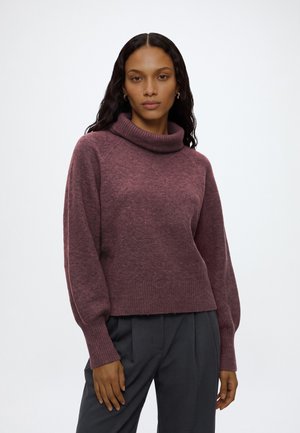 Femme aux longs cheveux foncés portant un pull à col roulé couleur prune et un pantalon noir, debout devant un fond clair uni.