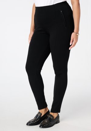 Vrouw in zwarte strakke broek met zijrits en zwarte gestudde loafers, staand met de handen ontspannen langs haar zij. 