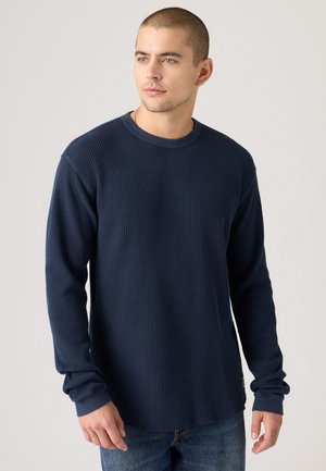 Chemise à manches longues bleu marine en texture gaufrée avec col rond et poignets côtelés, associée à un jean bleu clair.
