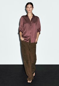 Camicia button-up in raso mauve con colletto e maniche arrotolate, abbinata a pantaloni in suede marrone a vita alta. Taglio semplice e rilassato.