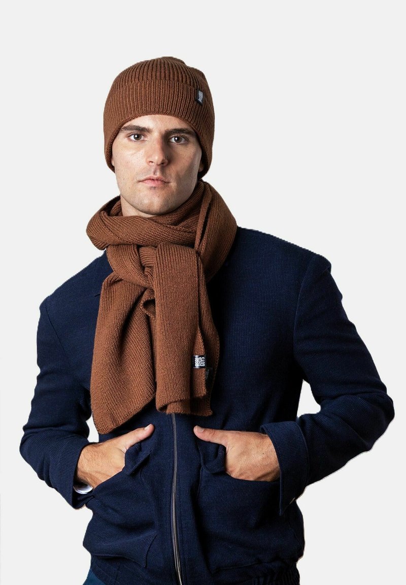 Mann trägt eine braune Strickmütze und dazu passenden Schal zu einer dunkelblauen Zip-Jacke, die Hände in den Taschen, vor einem schlichten hellen Hintergrund.