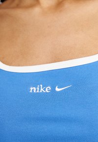 Mėlynas sportinis marškinėlis be rankovių su baltu apvadu, puošiamas išsiuvinėtu baltu Nike logotipu ant krūtinės. Lygus audinio paviršius.