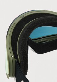 Detailný záber na zelené lyžiarske okuliare s modro sfarbeným sklom a hrubým čiernym penovým polstrovaním na bielom pozadí.