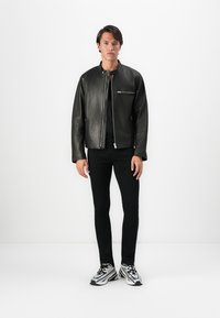Bărbat stând în fața unui fundal alb, îmbrăcat cu o jachetă de piele neagră, pantaloni negri și adidași argintii cu negru.