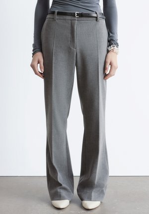 Person iført grå vidde bukser, sort bælte, grå langærmet top, hvide sko og sølvarmbånd, stående på grå gulv.