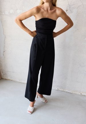 Frau trägt schwarzen trägerlosen Oberteil und schwarze weite Hosen, steht mit Händen in der Hüfte vor einer strukturierten hellen Wand.