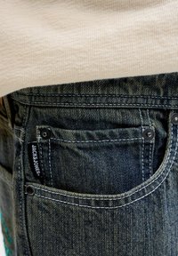 Κοντινό πλάνο στην πίσω τσέπη σκούρου τζιν με μαύρη ετικέτα Jack & Jones και μέρος από μπεζ πουλόβερ από πάνω.