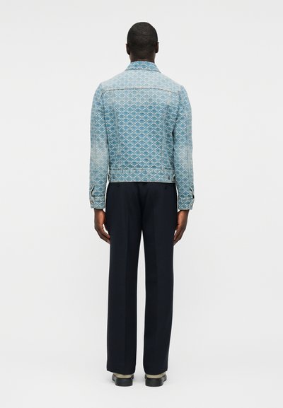 Άνδρας με πλάτη φοράει ένα ανοιχτό μπλε τζιν μπουφάν με σχέδια, μαύρο παντελόνι φαρδύ στο πόδι και μαύρα παπούτσια σε λευκό φόντο.