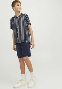 Chemise à rayures bleu marine et blanche à manches courtes avec des boutons, associée à un short bleu marine et des baskets blanches, se tenant sur un fond uni.