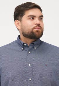 Koszula z guzikami w kolorze granatowym z geometrycznym wzorem. Posiada kołnierzyk, białe guziki oraz małe logo na piersi. Materiał bawełniany.