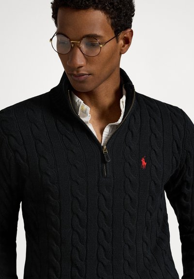 Μαύρο πουλόβερ με πλέξη καλώδιο, ψηλό κολάρο, φερμουάρ στο ένα τέταρτο, και μικρό κόκκινο λογότυπο πόλο στο στήθος. Υφή υφάσματος με ριμπ λεπτομέρειες.