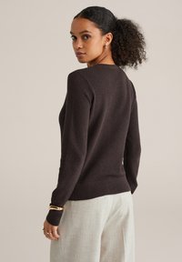 Rjavo teksturirano pleten pulover z dolgimi rokavi in okroglim izrezom, kombiniran s svetlimi hlačami po meri. Preprost rebrast rob.