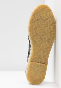 Chaussure noire avec une semelle en caoutchouc à motif, présentant une texture tissée. La semelle arbore une couleur beige clair avec un logo de taille visible.
