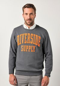 Uomo con barba che indossa un felpa grigia con la scritta arancione "RIVERSIDE SUPPLY", camicia bianca, pantaloni grigi chiari e un orologio al polso sinistro.