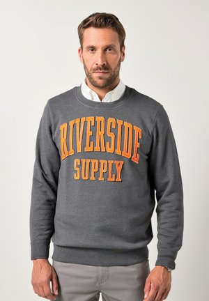 Man met een baard die een grijze sweatshirt draagt met oranje tekst "RIVERSIDE SUPPLY", een witte overhemden, lichtgrijze broek en een horloge om zijn linkse pols.