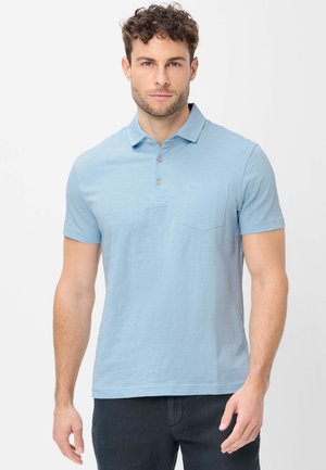 Mann mit lockigem Haar, der ein hellblaues kurzärmeliges Polohemd mit Brusttasche trägt, steht vor einem einfarbigen Hintergrund.