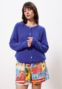 Femme aux cheveux bruns portant un cardigan bleu en peluche à boutons et un short à motifs colorés, se tenant devant un fond blanc.