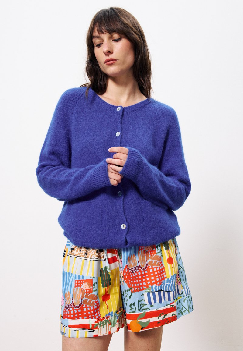 Femme aux cheveux bruns portant un cardigan bleu en peluche à boutons et un short à motifs colorés, se tenant devant un fond blanc.