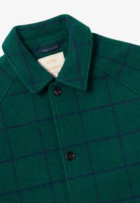 Grønn ullkåpe med blå rutemønster. Har krage, svart knappelukking og etikett inni. Myk tekstur, skreddersydd design.