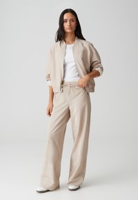 Veste bomber beige avec des coutures distinctives, assortie à un pantalon large, le tout en tissu lisse et léger. T-shirt blanc visible.