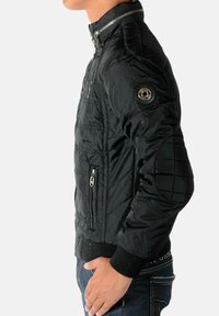 Sort quiltet jakke med opretstående krave, lynlås foran, ribstrikkede ærmegab og en logoaccent på venstre skulder. Glat, letvægtsstof.