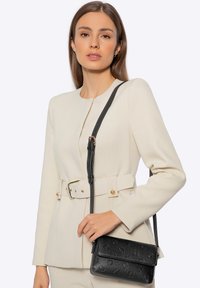 Beige getailleerd jasje met gouden knopen en een ceintuur in de taille, gecombineerd met een kleine zwarte crossbodytas met een textuurpatroon.