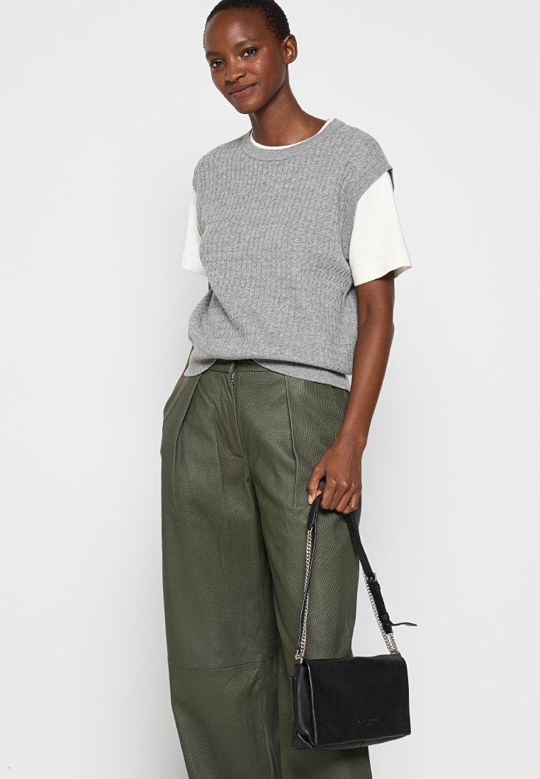 Grauer gestrickter ärmelloser Pullover über einem weißen Hemd, kombiniert mit lockeren grünen Lederhosen. Eine kleine schwarze Handtasche mit Kettenriemen wird gehalten.