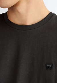 Nærbillede af en person iført en mørkegrå rundhals trøje med et lille sort trekantet logo med teksten "Wrangler" på brystet.