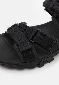 Svart utendørs sandal med justerbare nylonremmer, en polstret såle og teksturert gummisåle for godt grep.