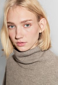 Maglione a collo alto in morbido lana beige, con un ampio colletto a coste e una vestibilità rilassata, indossato da una persona con capelli biondi chiari.