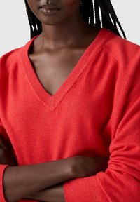 Pull rouge en V avec une texture douce, comportant des poignets côtelés et une coupe décontractée, présentant un léger motif chiné sur le tissu.